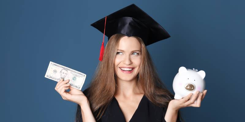 Graduate holding a dollar bill in one hand and a piggy bank in the other