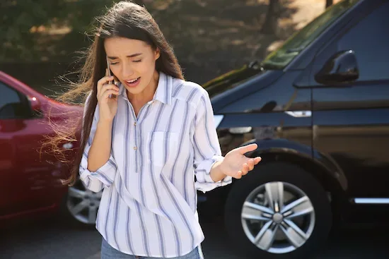 Woman calling for help after a car accident, standing next to her car talking on a cell phone.